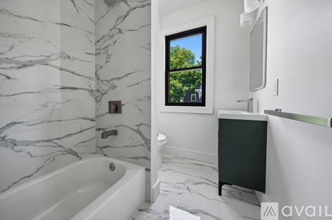 A bathroom with a marble patterned floor and walls, a white bathtub, a window with a view of trees, and a green cabinet.