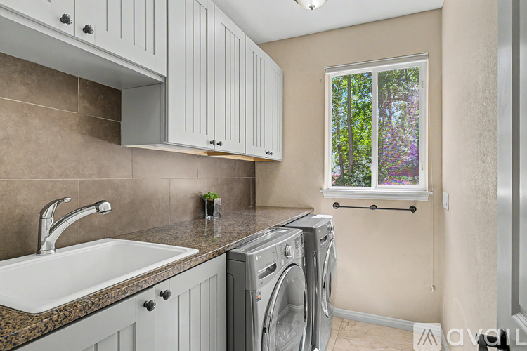 A modern laundry room with a washer and dryer, a sink, and a window with a view of trees.