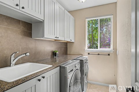 A modern laundry room with a washer and dryer, a sink, and a window with a view of trees.