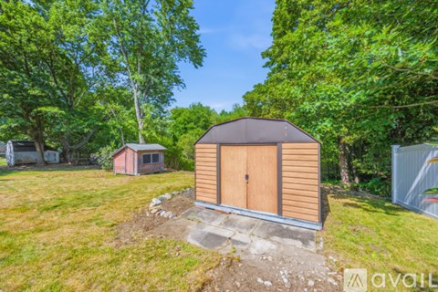 A wooden shed with a brown roof is situated in a grassy area with trees in the background.