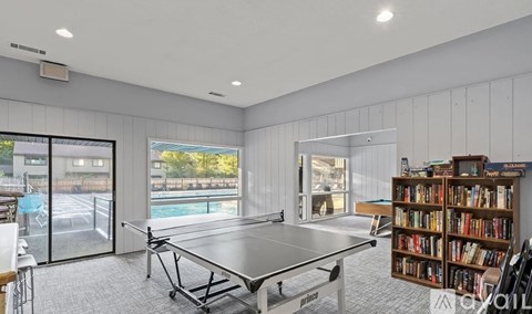 A room with a ping pong table, a bookshelf, and a view of a pool outside.