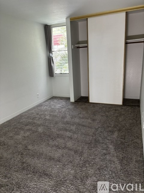 A room with grey carpet and a window with a curtain.