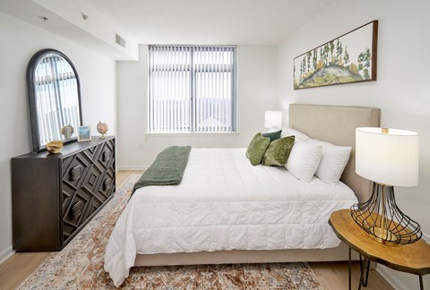 A bedroom with a large bed, a side table with a lamp, and a rug on the floor.