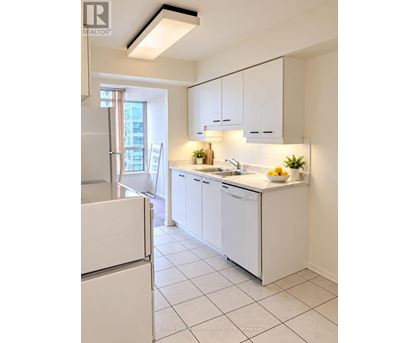 A white kitchen with a refrigerator on the left and a sink with a bowl of fruit on the counter.