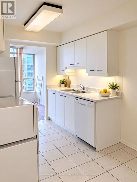 A white kitchen with a refrigerator on the left and a sink with a bowl of fruit on the counter.