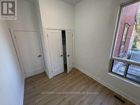 A room with white walls and wooden flooring, with a window showing a red building outside.