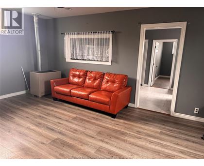 A red leather couch sits in a room with wooden flooring and a window with white curtains.