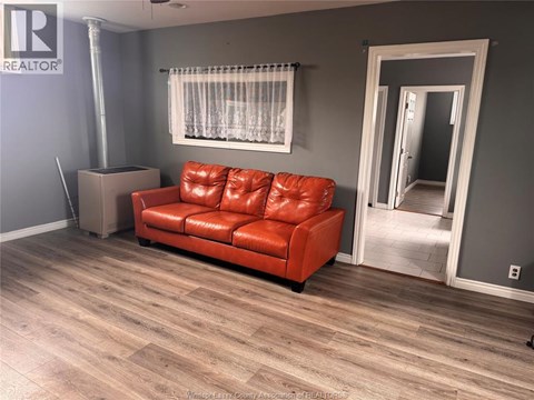 A red leather couch sits in a room with wooden flooring and a window with white curtains.