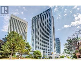A tall building with a glass facade stands in front of a smaller building and trees.