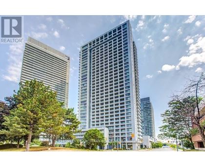 A tall building with a glass facade stands in front of a smaller building and trees.