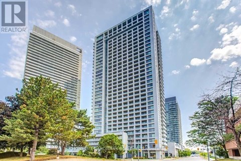 A tall building with a glass facade stands in front of a smaller building and trees.