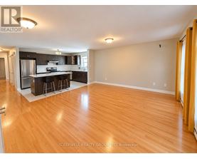 A well-lit, empty living room with wooden flooring and a kitchen area in the background.