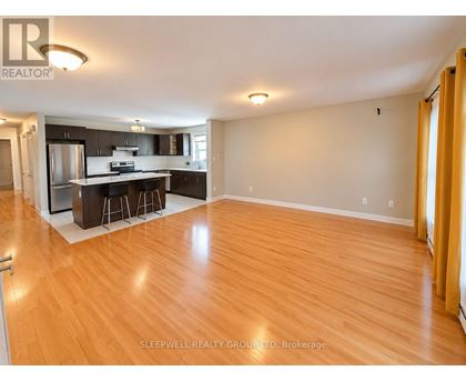 A well-lit, empty living room with wooden flooring and a kitchen area in the background.