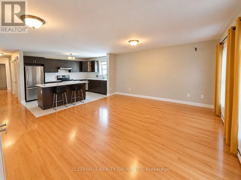 A well-lit, empty living room with wooden flooring and a kitchen area in the background.