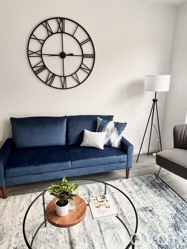 A living room with a blue couch, a round clock on the wall, and a small potted plant on a coffee table.