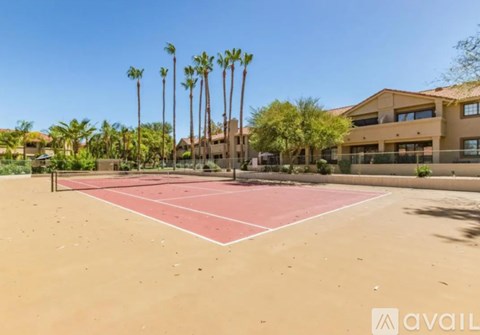 A tennis court with a red surface surrounded by palm trees and a building in the background.