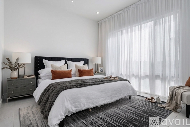 A bedroom with a large bed, a nightstand with a plant, and a window with sheer curtains.
