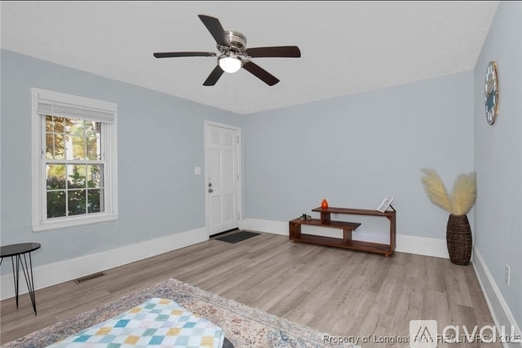 A room with a ceiling fan, a window, a door, a bench, a table, and a vase on the floor.