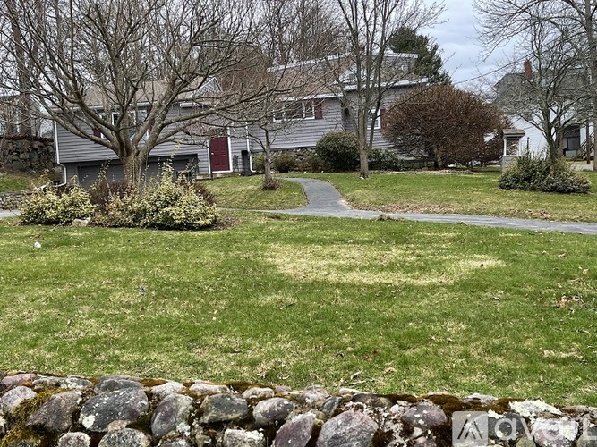 A house with a red door is surrounded by a grassy lawn and a stone wall.