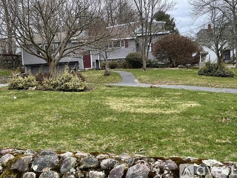 A house with a red door is surrounded by a grassy lawn and a stone wall.