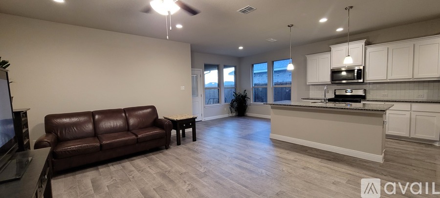 A living room with a brown leather couch and a kitchen area with white cabinets.