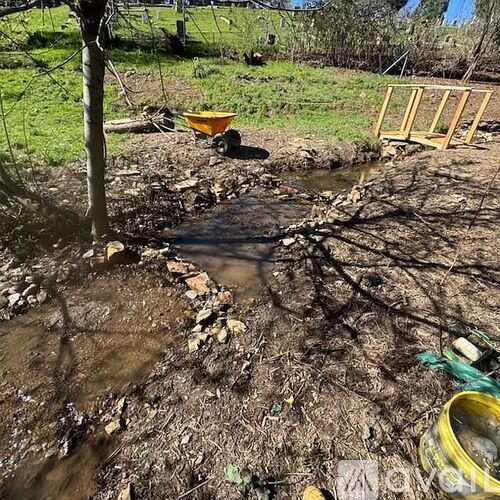 A muddy stream flows through a grassy area with a yellow wheelbarrow and wooden structure nearby.
