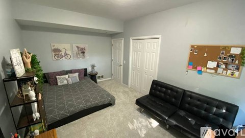 A black leather sofa sits in a living room with a bed and a cork board on the wall.