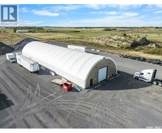 A large white building with a curved roof is surrounded by trucks and a red container on a dirt ground.