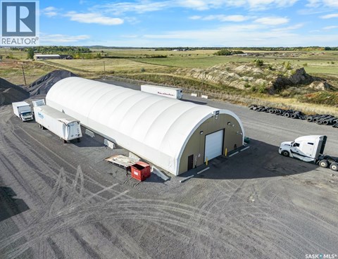 A large white building with a curved roof is surrounded by trucks and a red container on a dirt ground.