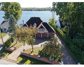 A house with a black roof and white walls is surrounded by trees and a body of water.