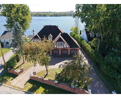 A house with a black roof and white walls is surrounded by trees and a body of water.