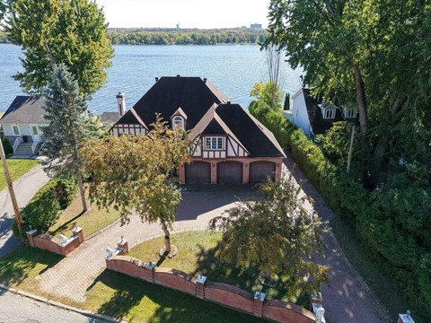 A house with a black roof and white walls is surrounded by trees and a body of water.