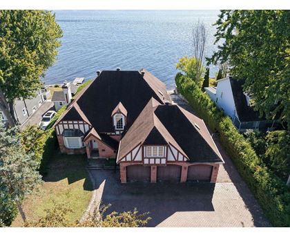 A house with a brown roof and white walls is surrounded by a brick driveway and green trees.