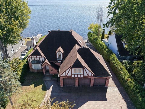 A house with a brown roof and white walls is surrounded by a brick driveway and green trees.
