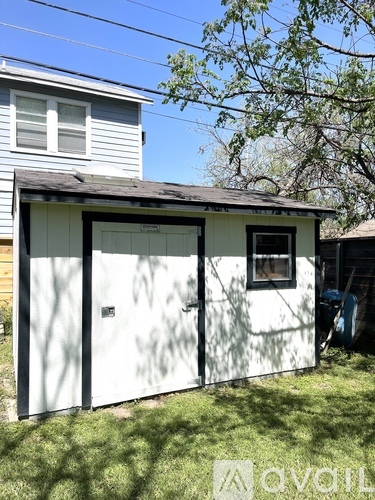 A small white shed with a black door and a window is situated in a grassy area.