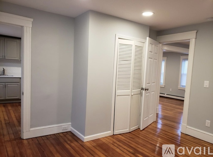 A room with wooden floors and white walls, with a kitchen visible through an open door.