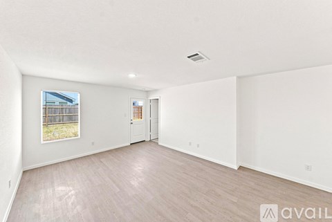 A spacious room with light wood flooring and white walls, with a window showing an outdoor fence and trees.