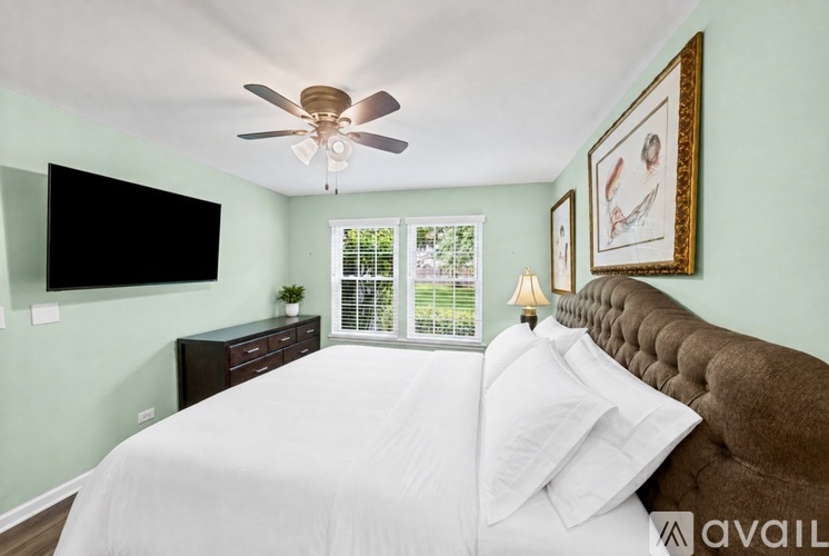 A bedroom with a brown headboard bed, a white bedspread, a ceiling fan, a black flat screen TV, a window with blinds, a nightstand with a lamp and a plant, and two framed pictures on the