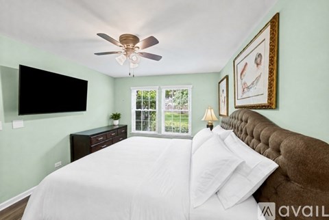 A bedroom with a brown headboard bed, a white bedspread, a ceiling fan, a black flat screen TV, a window with blinds, a nightstand with a lamp and a plant, and two framed pictures on the