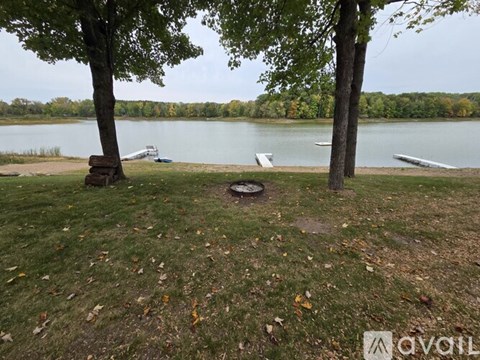 A tranquil scene of a lake with boats docked on the shore, surrounded by trees and a grassy area with fallen leaves.