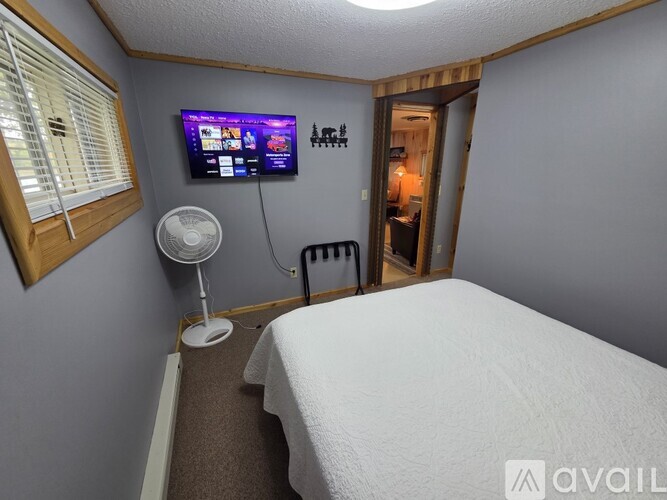 A small bedroom with a bed, a fan, a window with blinds, a door, and a TV mounted on the wall.