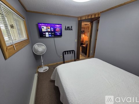 A small bedroom with a bed, a fan, a window with blinds, a door, and a TV mounted on the wall.