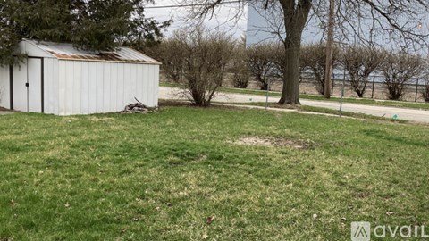 A white shed sits in a grassy field with a tree and a fence in the background.