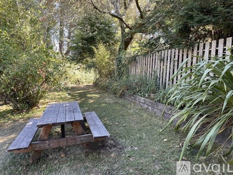 A wooden picnic table sits in a grassy area surrounded by trees and a wooden fence.