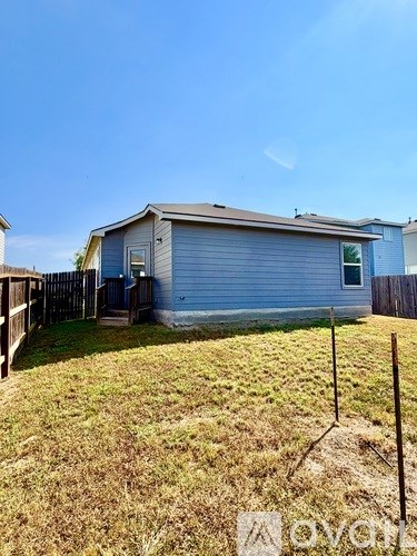 A blue house with a white door and windows is surrounded by a fence and has a grassy yard.