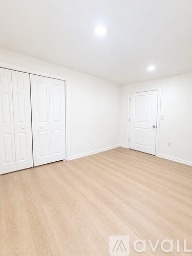 A room with white walls and wooden flooring, with a white door on the right and a white wardrobe on the left.