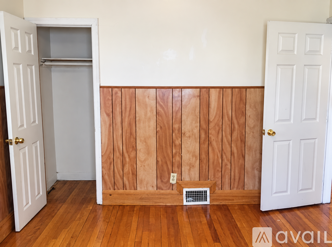 A room with a white door, a white wall, and a wooden floor with a vent.