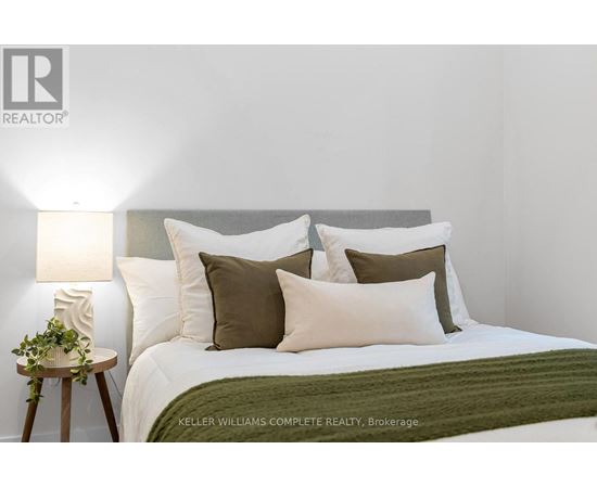 A bedroom with a bed, pillows, a lamp, and a small table with a plant on it.