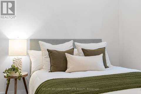 A bedroom with a bed, pillows, a lamp, and a small table with a plant on it.