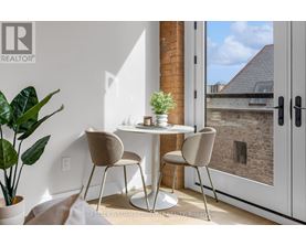 A table with two chairs and a potted plant sits in a room with a view of a brick building outside.